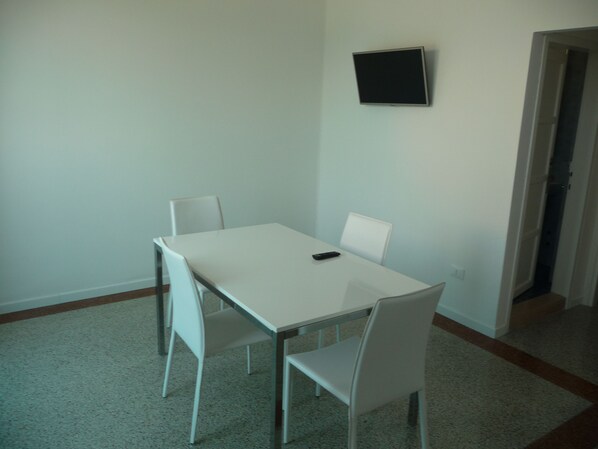 A small, sparsely furnished room with a white table and two white chairs, a remote control on the table, and a flat-screen TV mounted on the wall. - Soggiorno Aurora (Caorle)