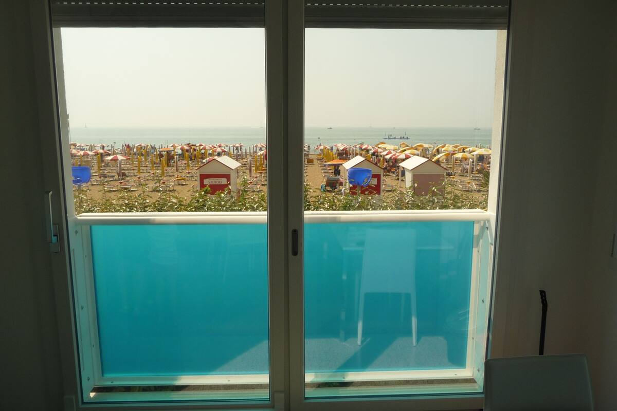 Une pièce dotée d’une grande fenêtre donnant sur une plage parsemée de parasols et de cabanes de plage.
