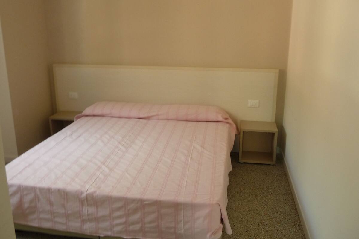 Une petite chambre avec un lit simple, une couverture unie et deux tables de chevet.