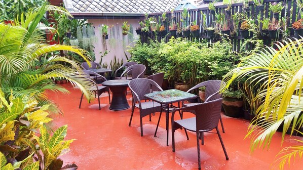 An outdoor patio area with wicker chairs and tables, surrounded by potted plants and a red floor. - PAKHONGTHONG VILLA (Luang Prabang)