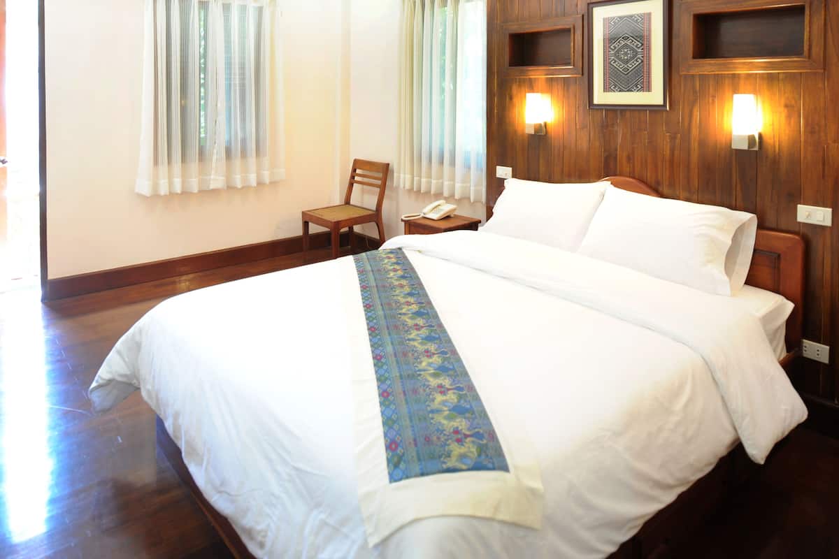 A hotel room with a large bed, wooden headboard, a small wooden chair, a bedside table with a lamp, and a wall-mounted phone.