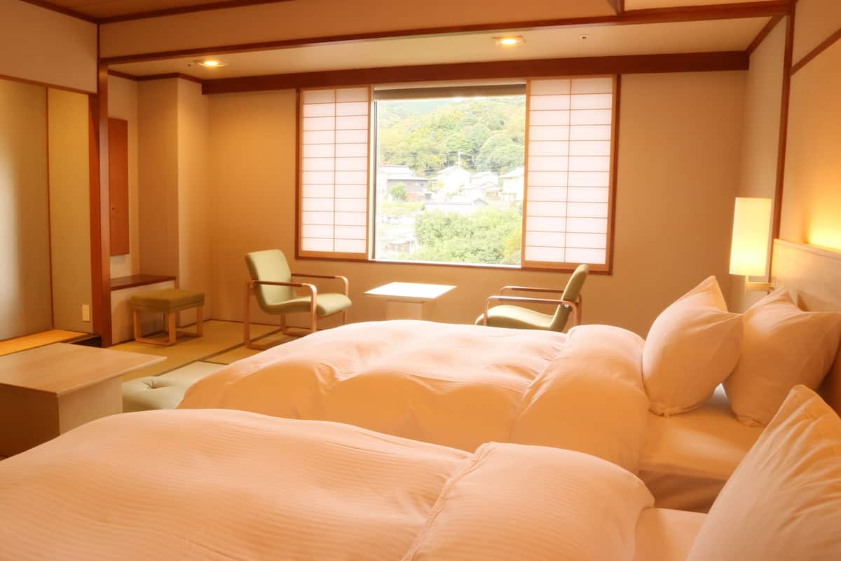 A traditional Japanese-style room with tatami flooring, a low table, a chair, a bed with white bedding, and a window with shoji screens.