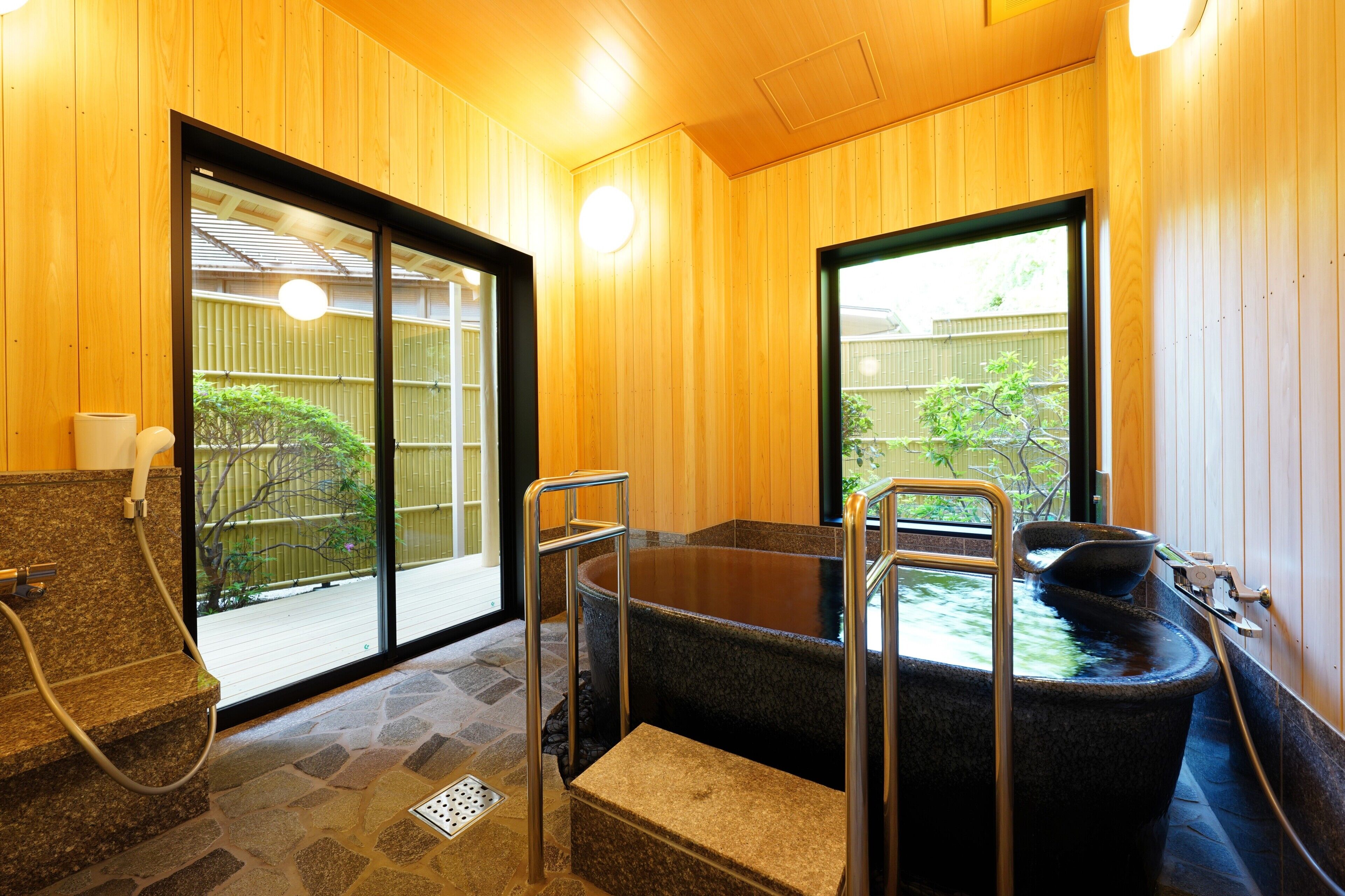 A traditional Japanese onsen with a stone hot spring, wooden walls, and a view of a bamboo fence outside.