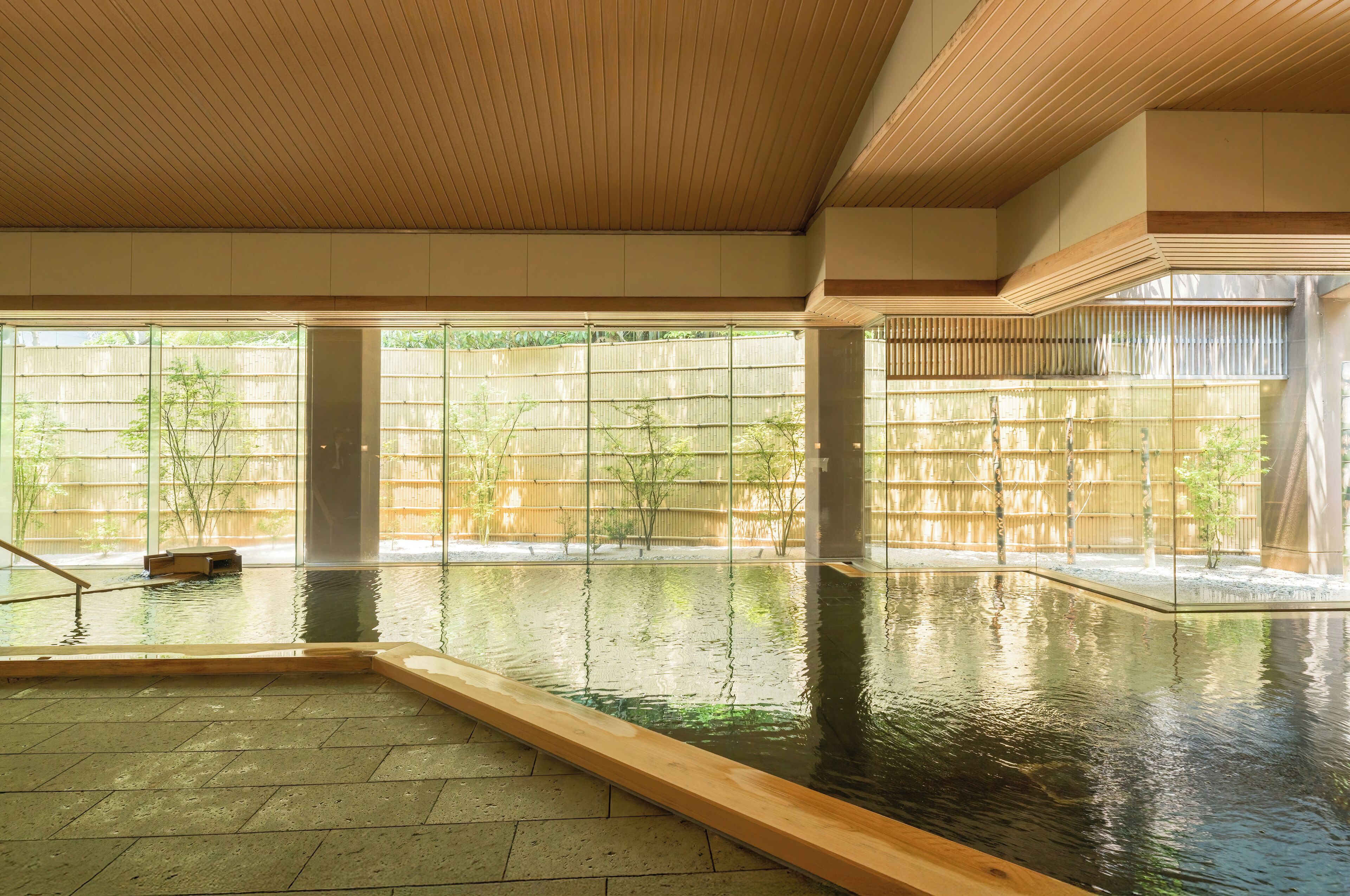 Una piscina interior con paredes de vidrio transparente, suelo de madera y una valla de bambú visible en el exterior.