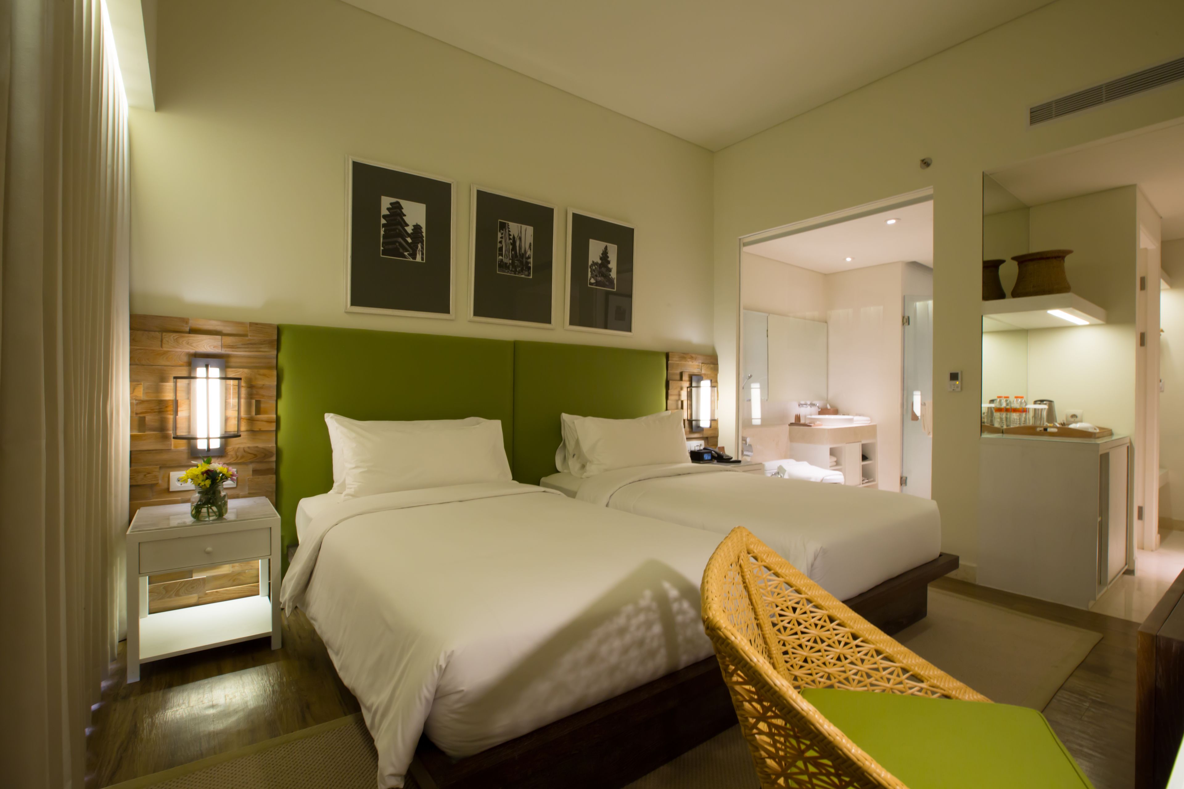 A hotel room with two beds, a green headboard, a nightstand with a lamp, and a wicker chair.