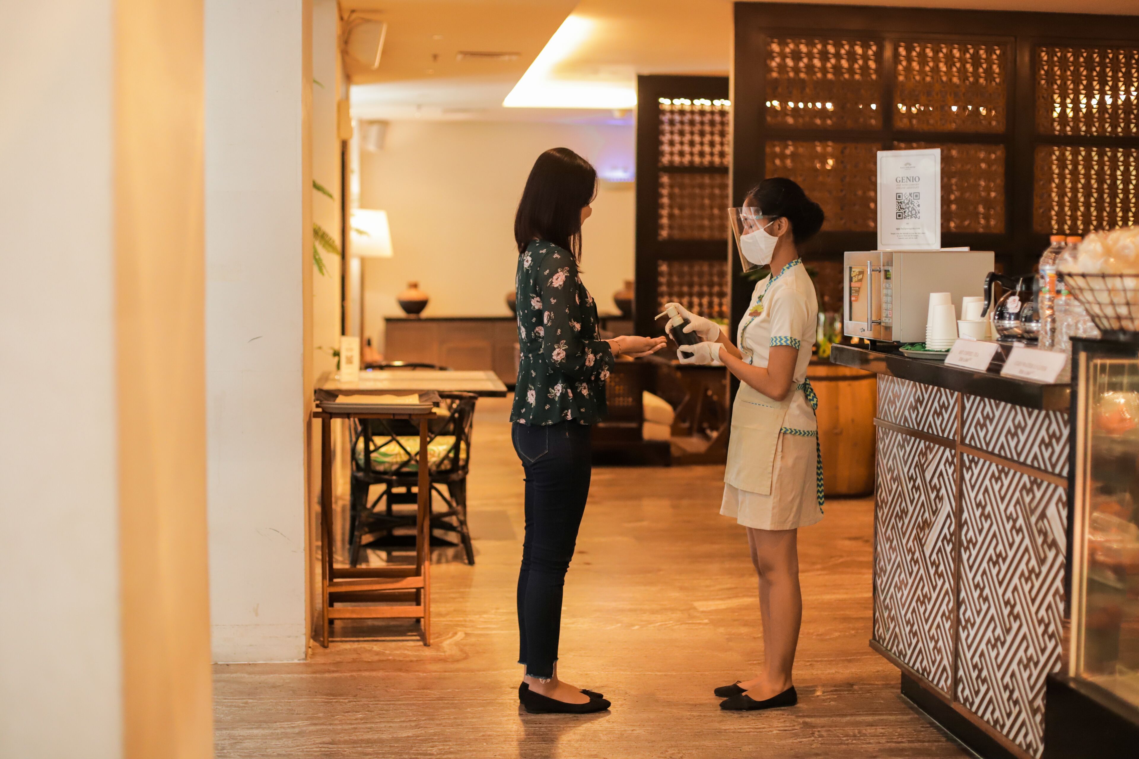 A woman in a floral shirt and jeans standing at a counter in a restaurant with a patterned wall and a QR code sign.