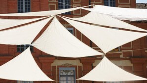 Une série de toiles triangulaires blanches, disposées selon un motif géométrique, se détachent sur le mur d’un bâtiment en briques agrémenté d’une fenêtre décorative.