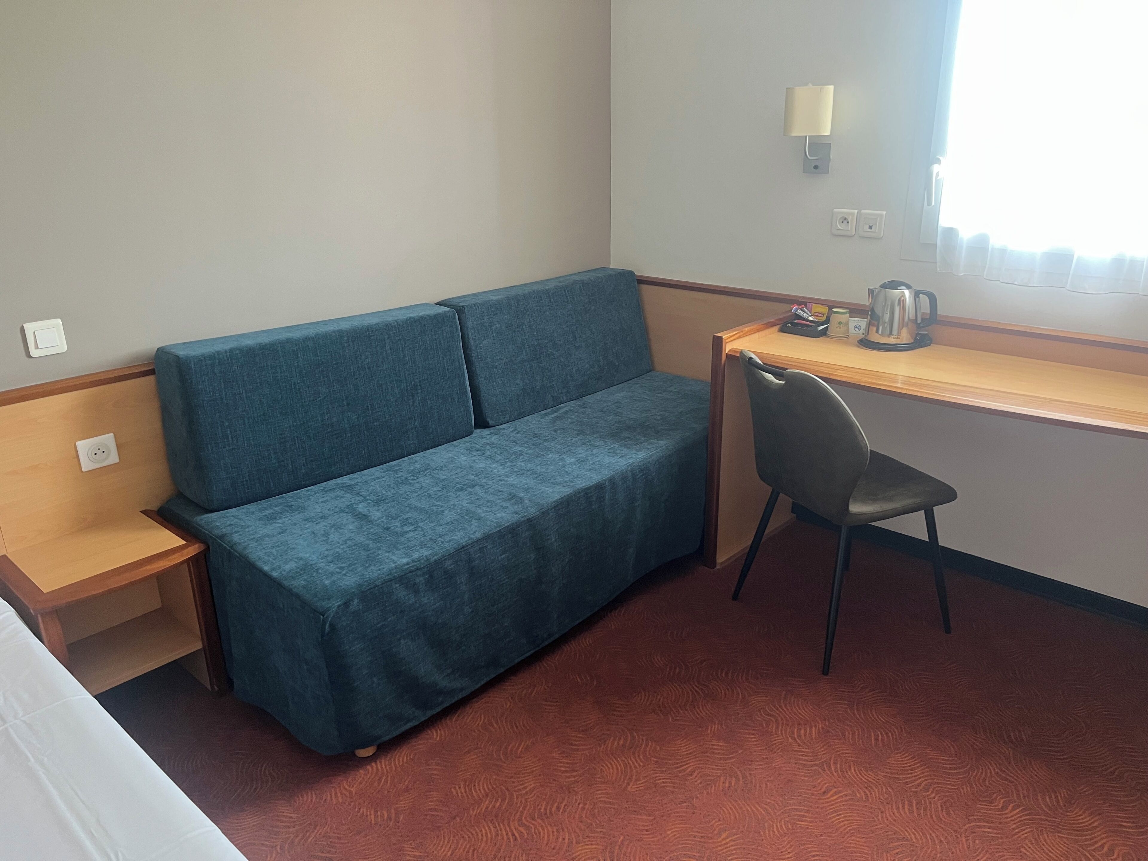 A small room with a blue sofa, a wooden desk, a black chair, and a window with curtains.