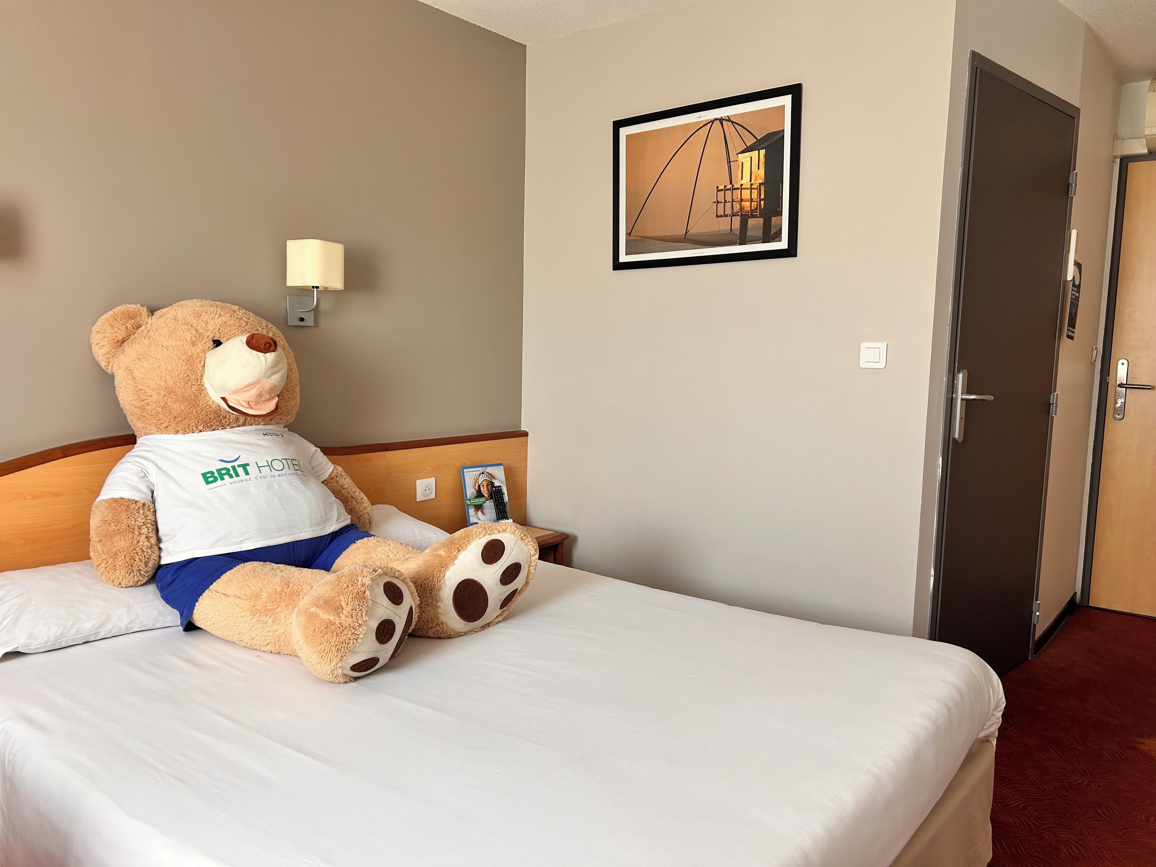 Une chambre d’hôtel avec un grand ours en peluche sur le lit, un tableau encadré au mur et une porte sur le côté droit.