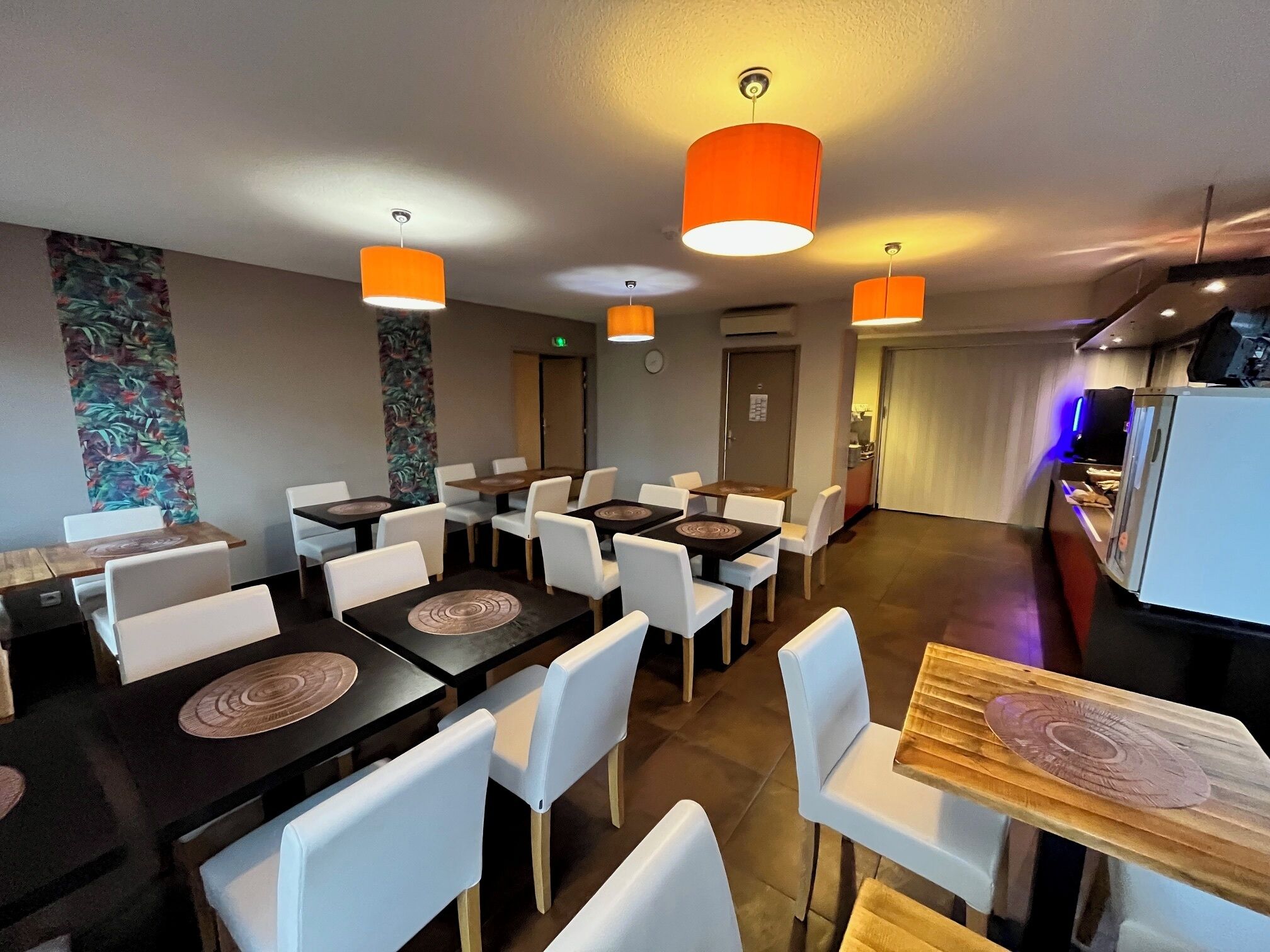 A dining area with tables and chairs, orange pendant lights, and a kitchen area with a refrigerator.