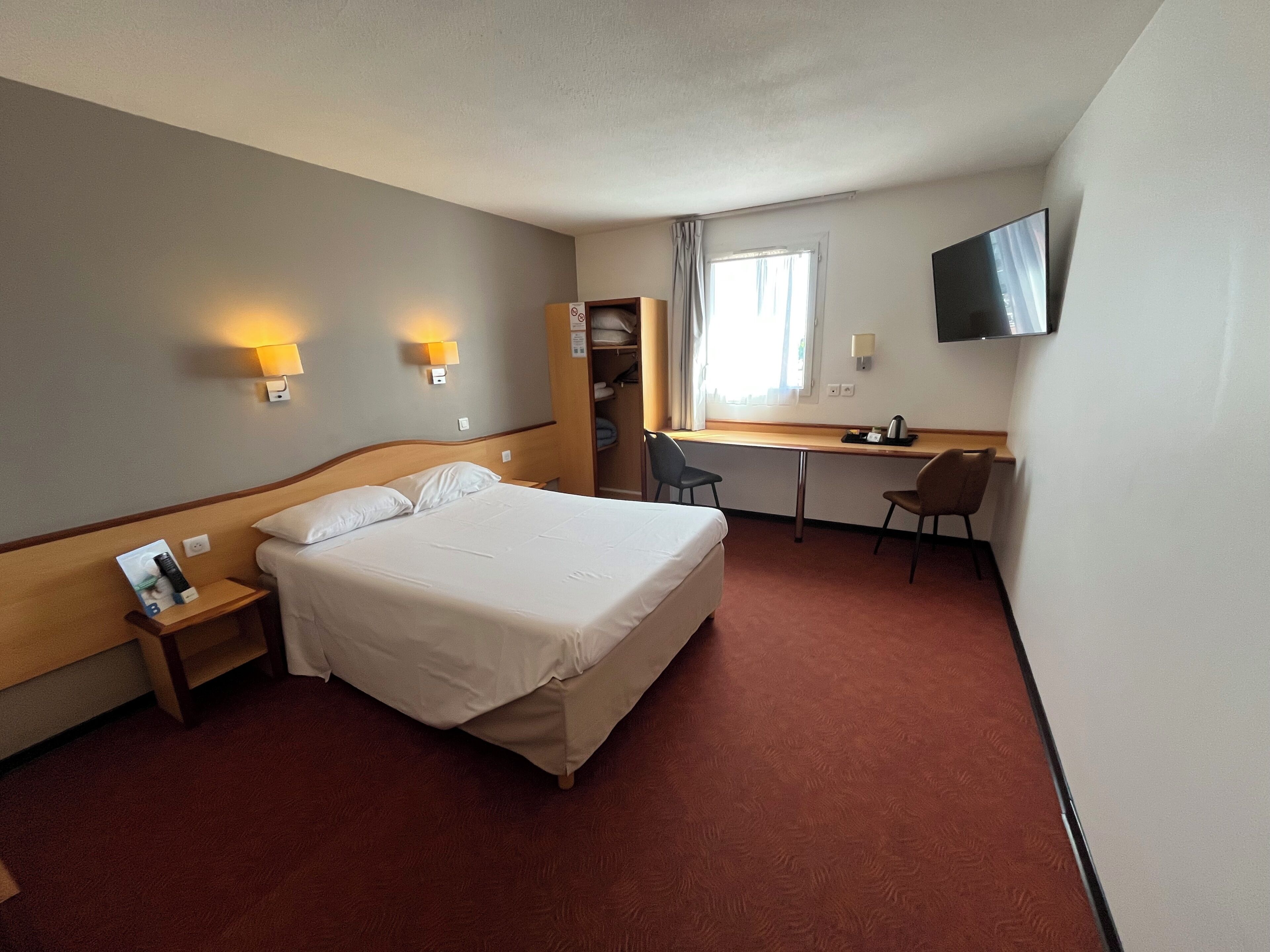 A hotel room with a bed, desk, chair, TV, and a wardrobe.