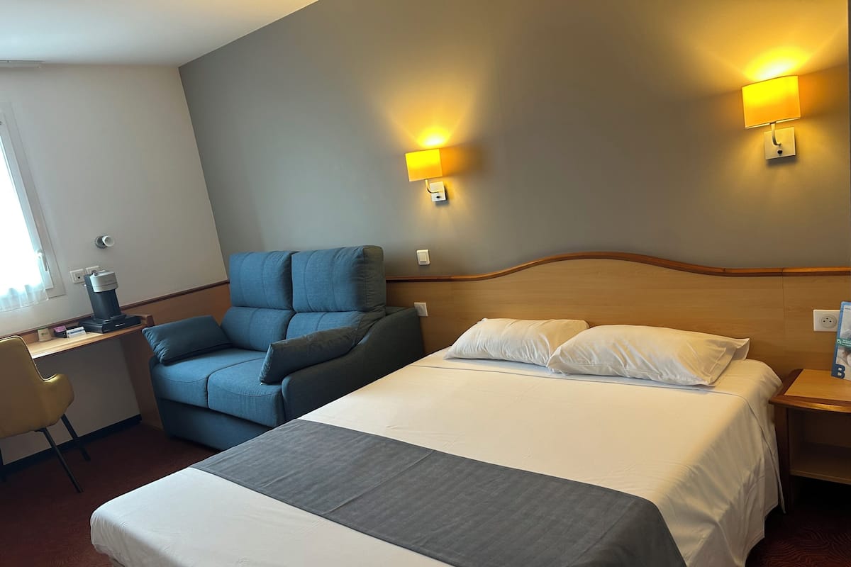 Een hotelkamer met een groot bed, een bureau met stoel, een blauwe fauteuil en twee wandlampen.