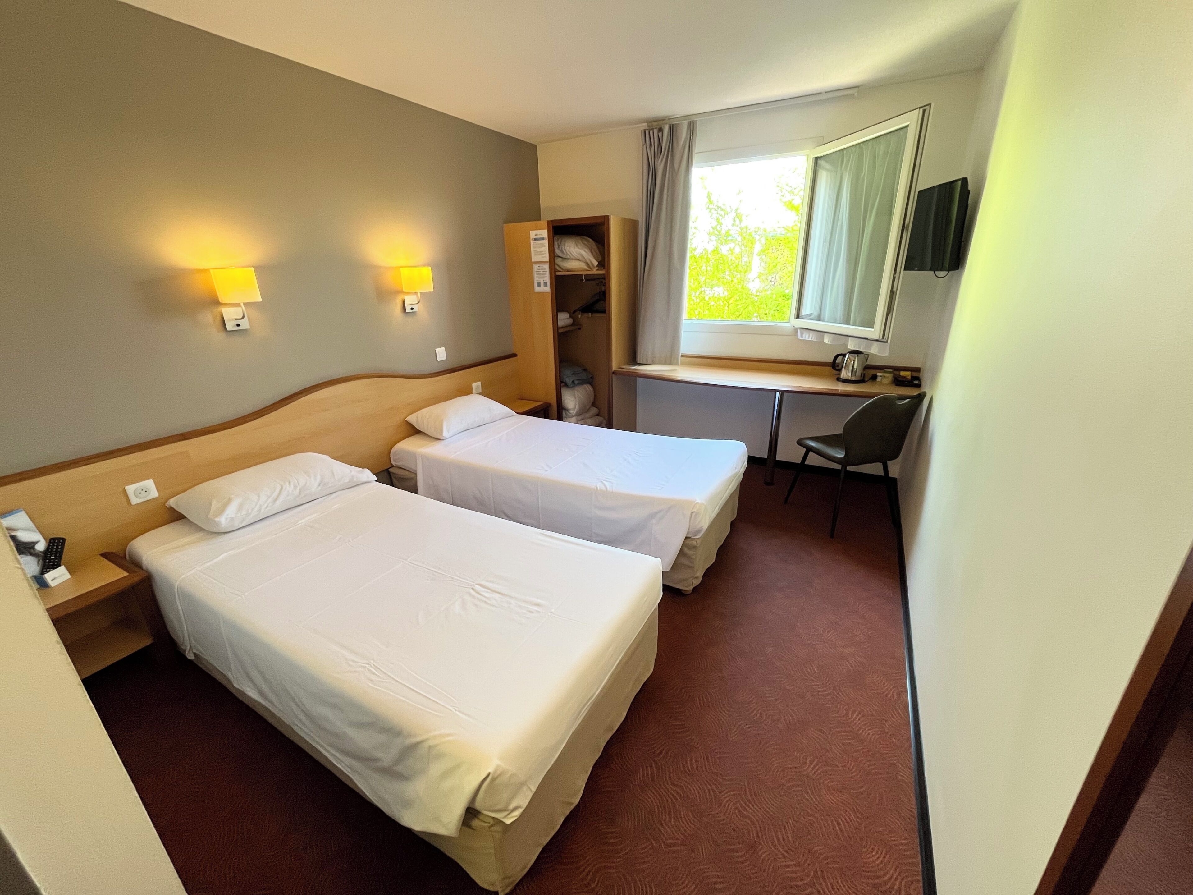 A hotel room with two beds, a desk, a chair, a TV, and a window with curtains.