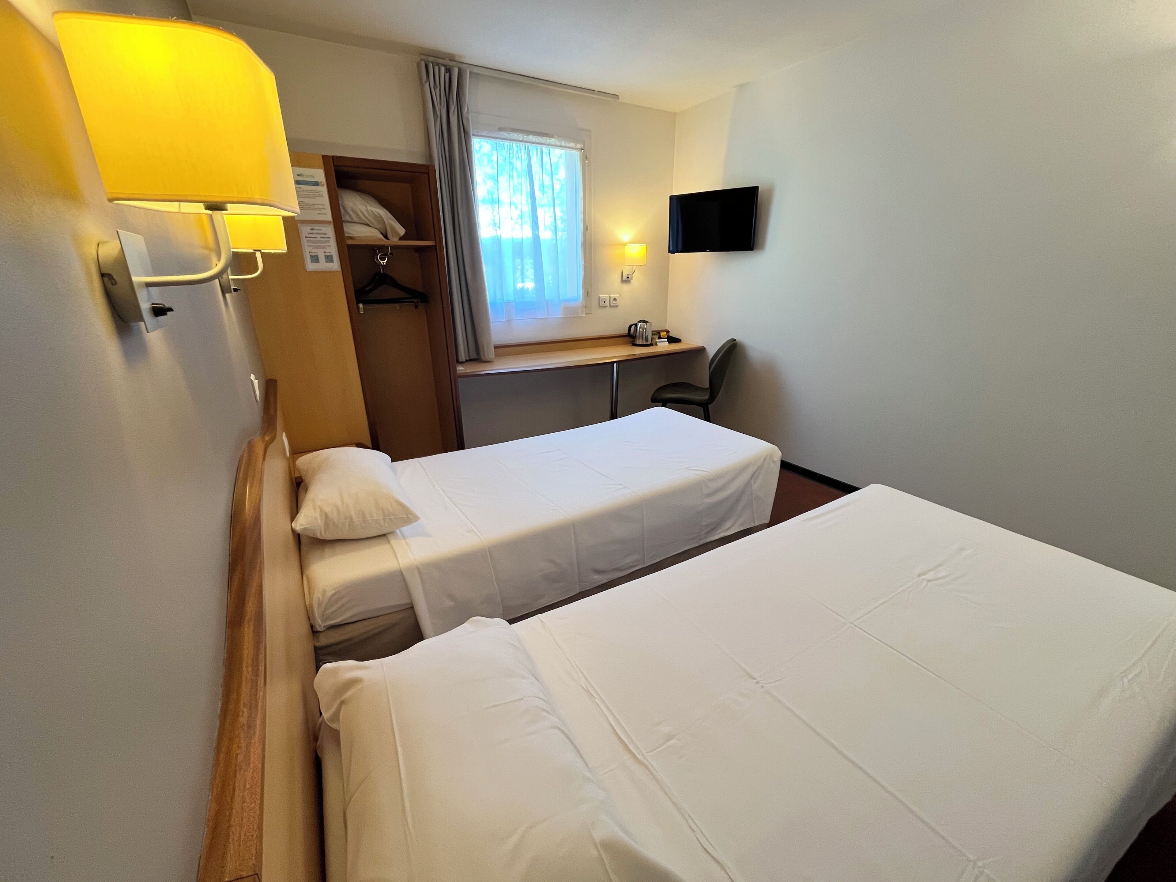 A hotel room with two beds, a desk, a chair, a TV, and a window with curtains.