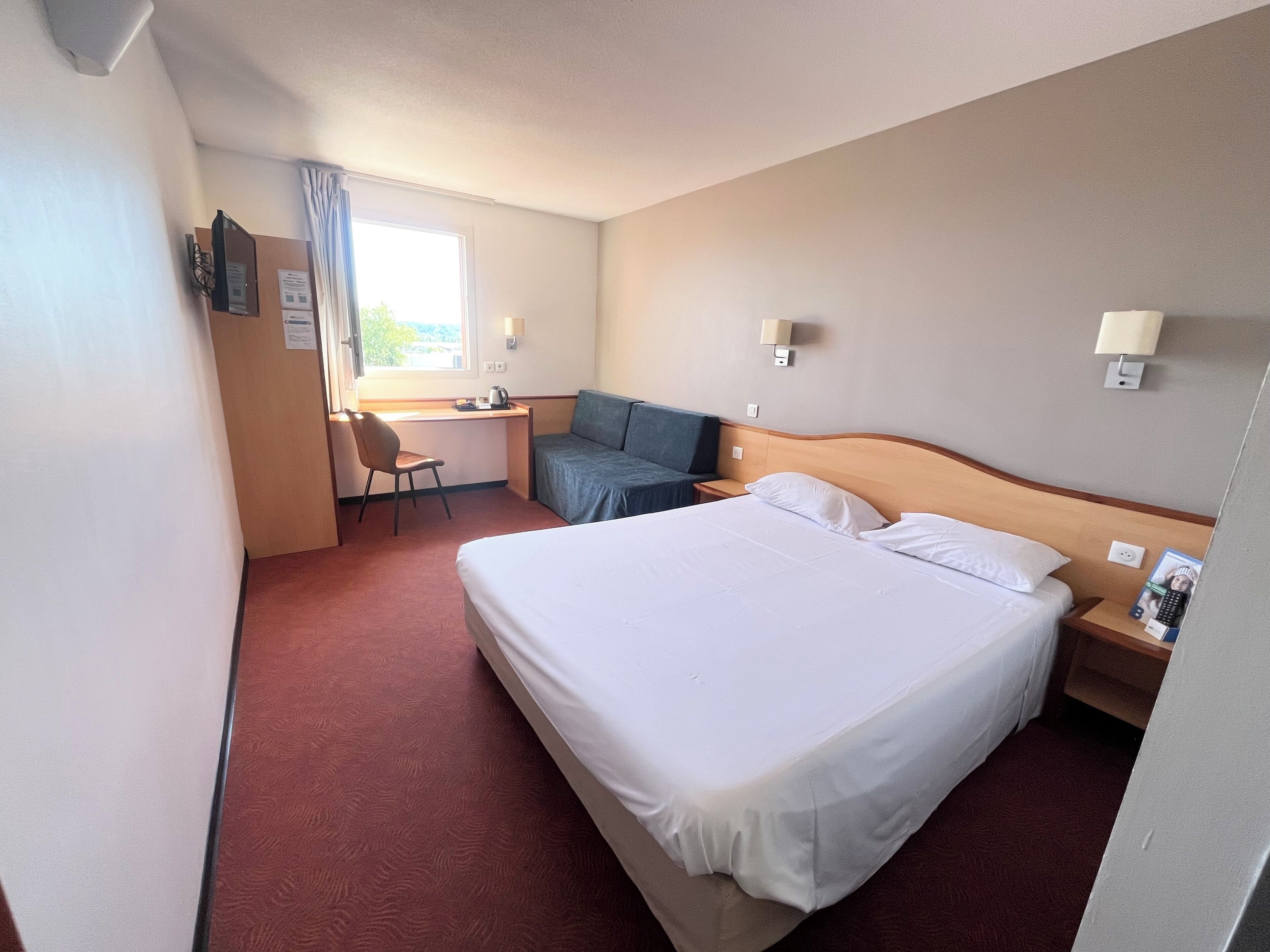 A hotel room with a bed, desk, chair, and a small window.