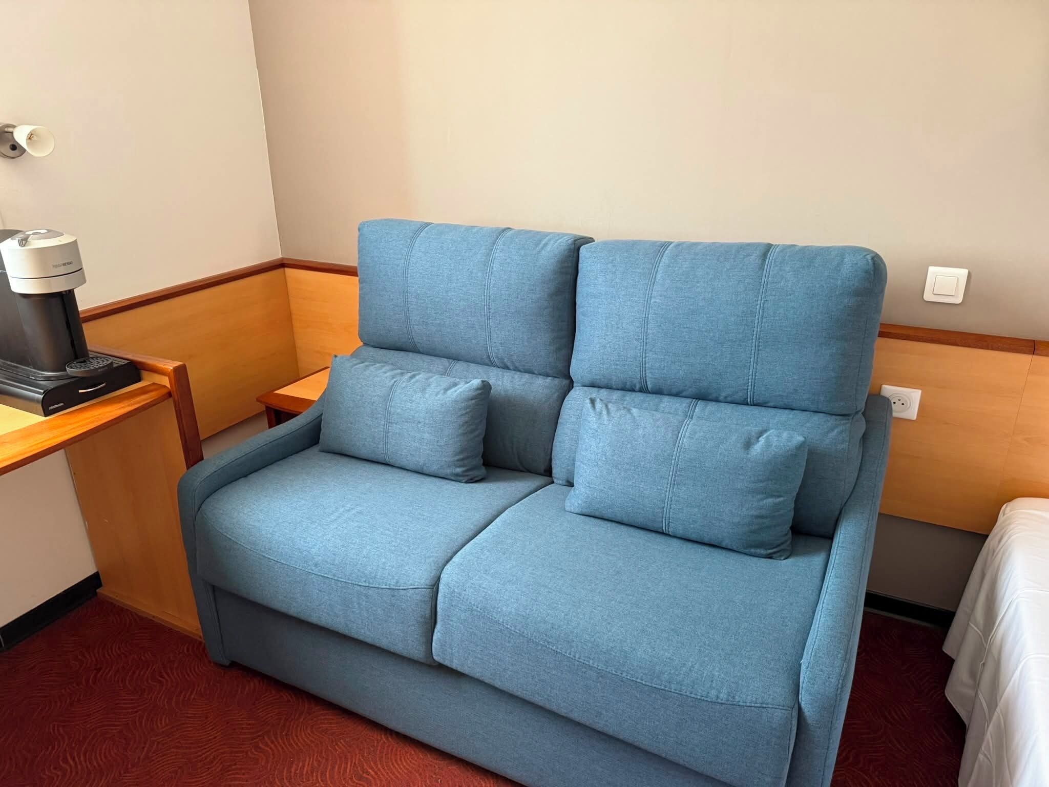 Un sofá azul con dos almohadas, una cafetera sobre una mesa de madera y una cama con ropa blanca.