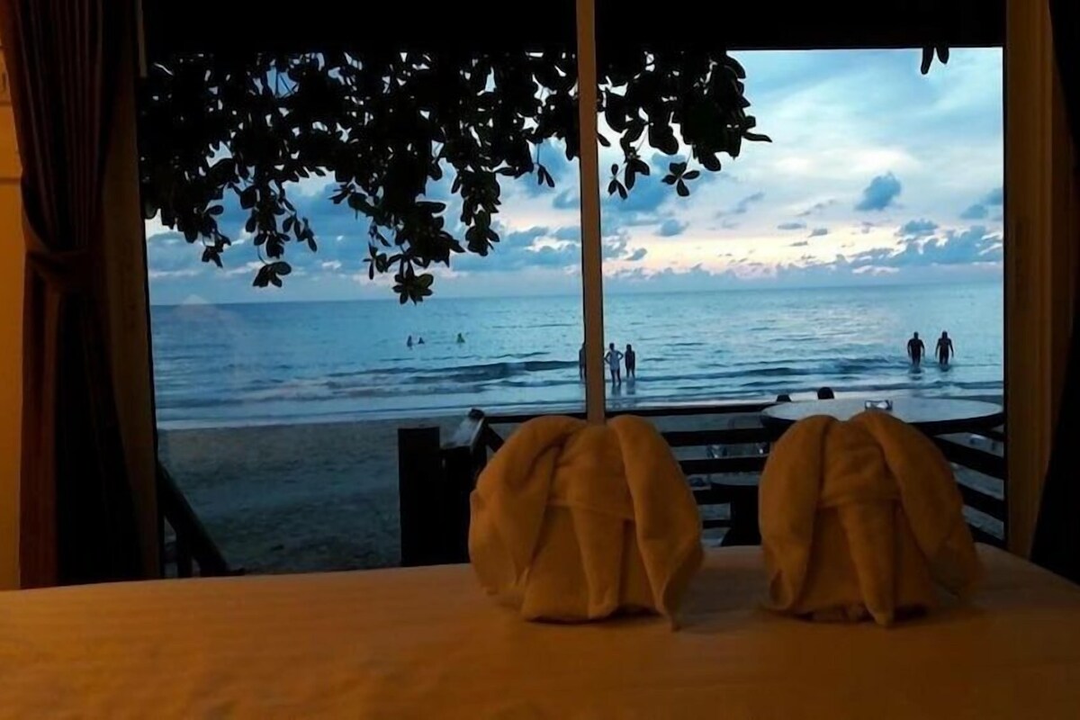 Una habitación con vista a la playa, una cama con cubrecama blanco y una ventana con cortinas.