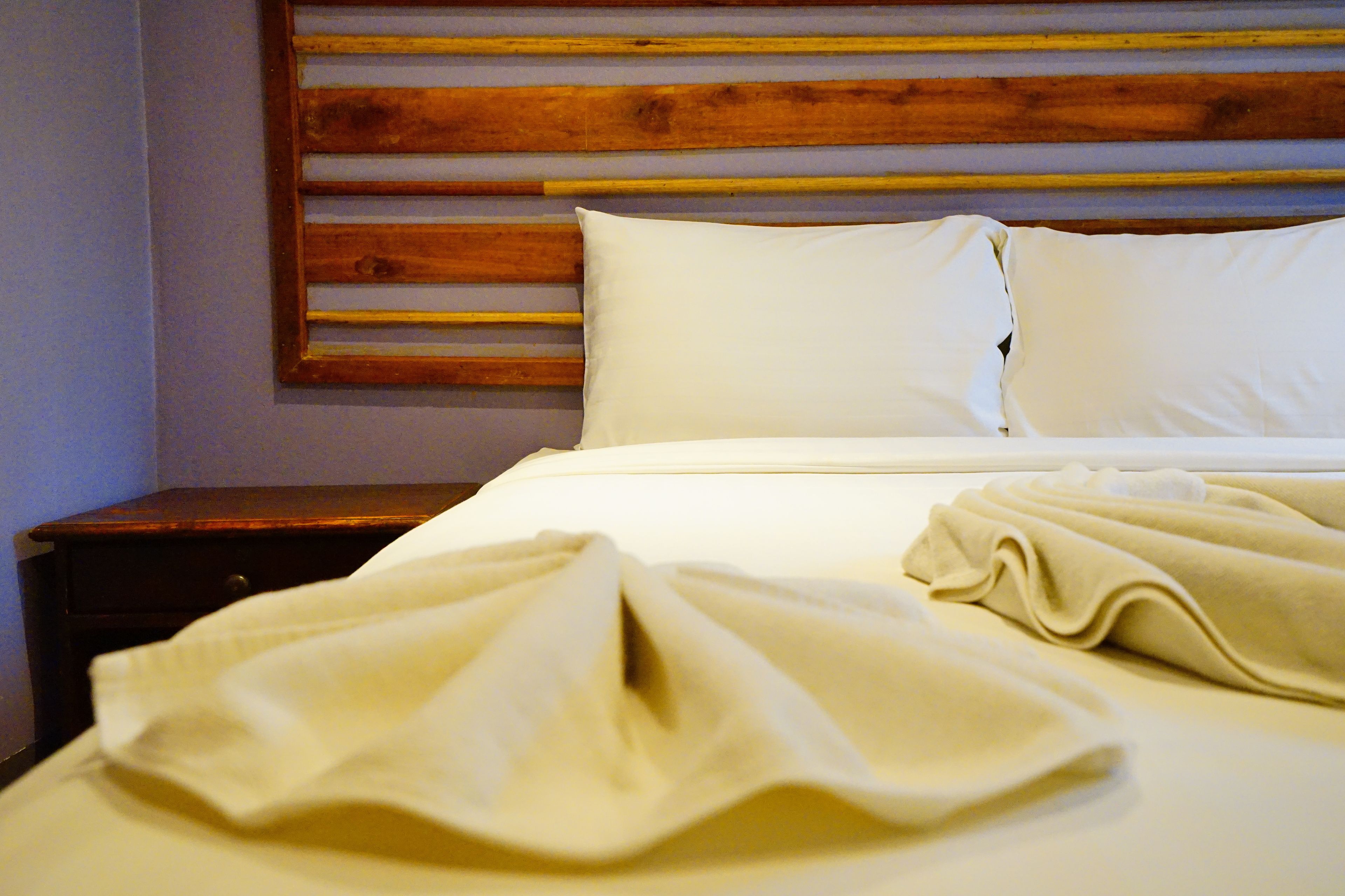 Ein ordentlich gemachtes Bett mit weißen Bettwäsche und Kissen, ein hölzernes Kopfteil und ein kleiner Nachttisch daneben.