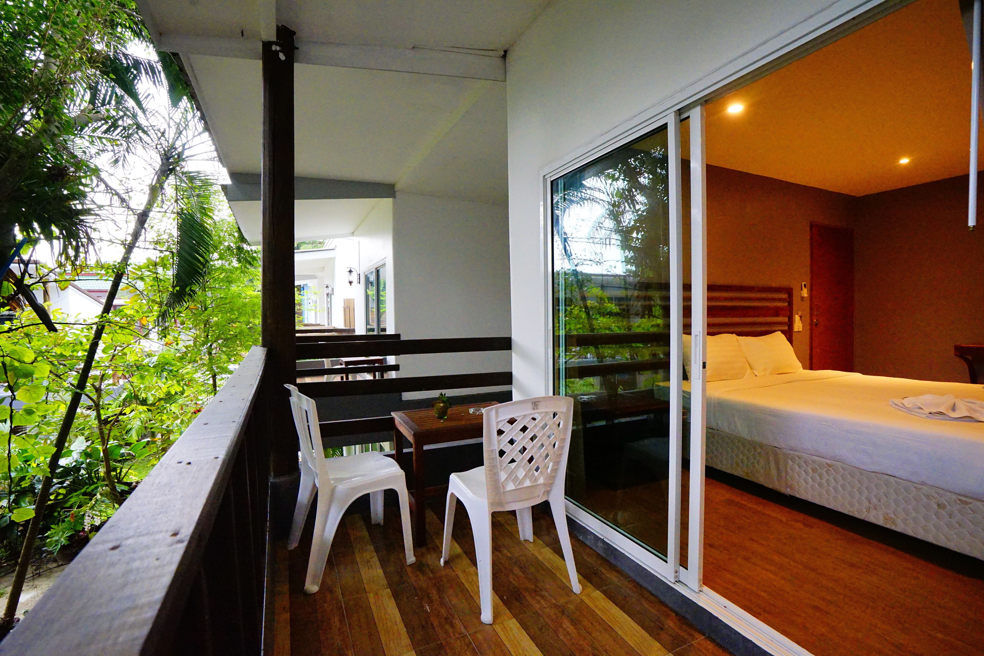 A balcony with a wooden table, two white chairs, and a glass door leading to a bedroom with a bed and a nightstand.