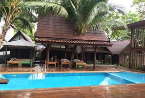 Eine Holzterrasse mit Pool, Palmen und einer überdachten Sitzgelegenheit mit einer Person.