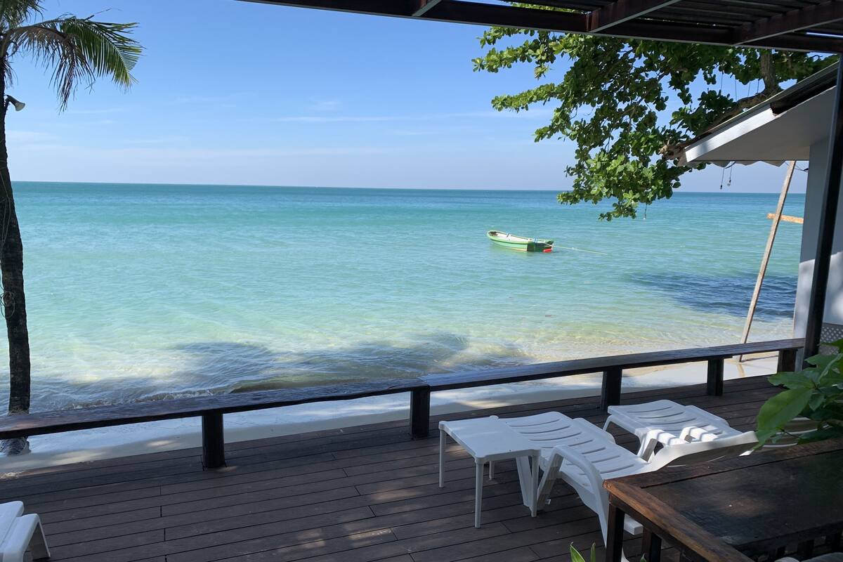 Eine Holzterrasse mit weißen Kunststoffstühlen und einem Tisch, mit Blick auf das klare, türkisfarbene Meer, wo ein Boot vor Anker liegt.