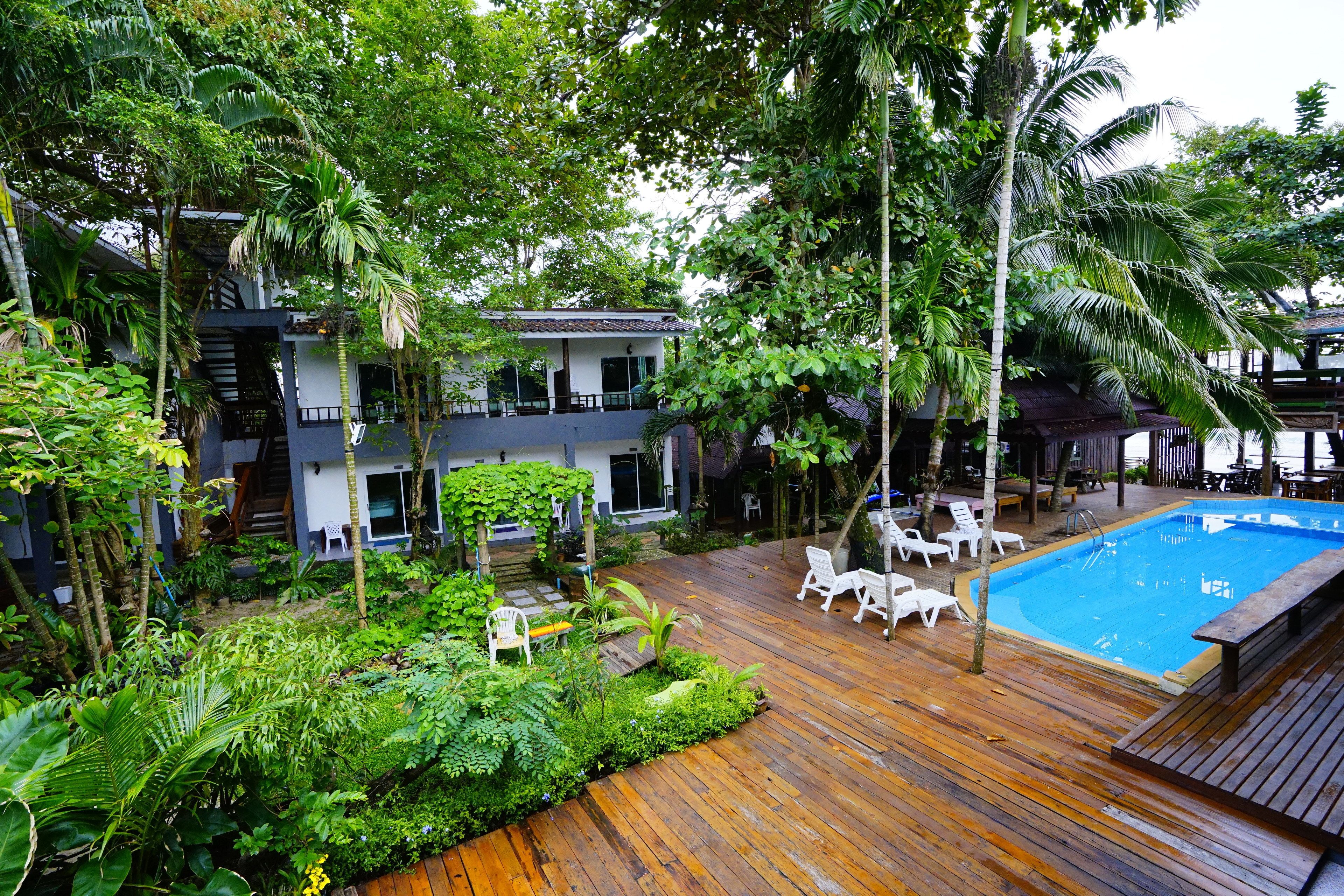 Una terraza de madera con piscina, rodeada de exuberante follaje y sillones blancos.