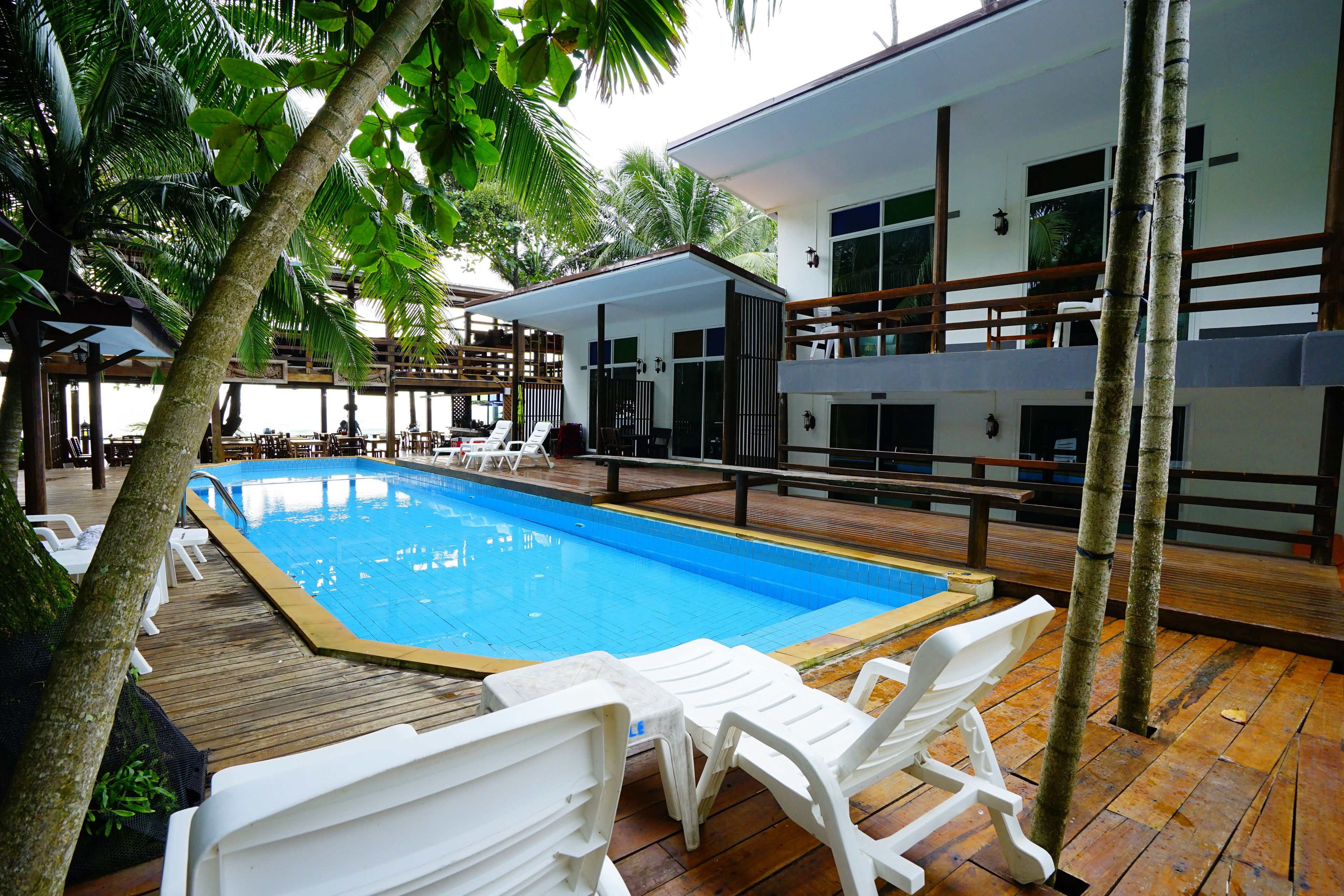 Una piscina con sillas de descanso blancas, una terraza de madera y un edificio con balcones.