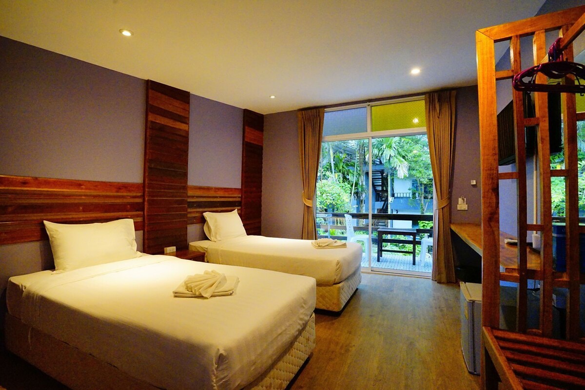 A hotel room with two beds, a wooden headboard, a balcony with a table, and a view of greenery.