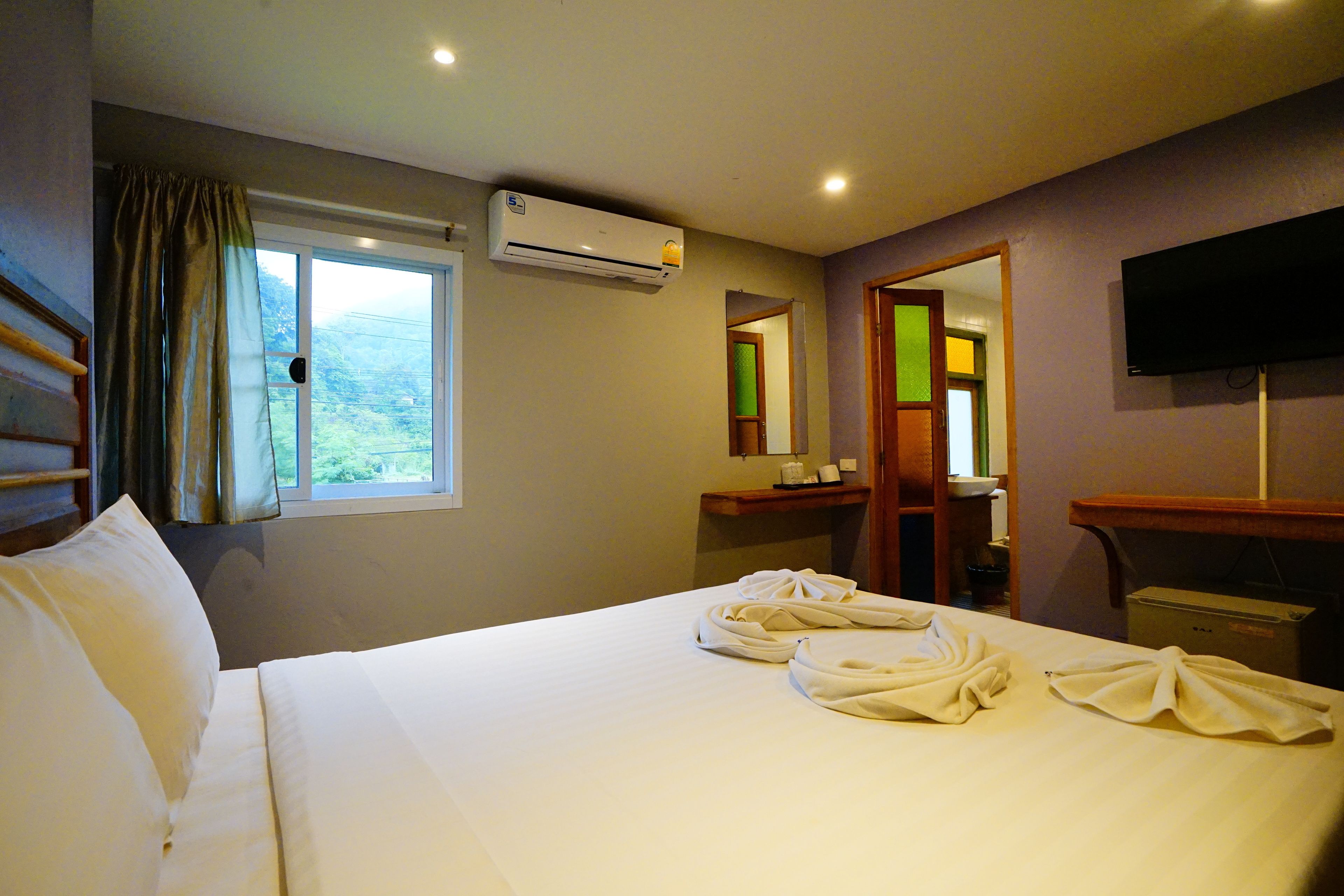 Quarto de hotel com cama, janela, ar condicionado, espelho, televisão e banheiro.