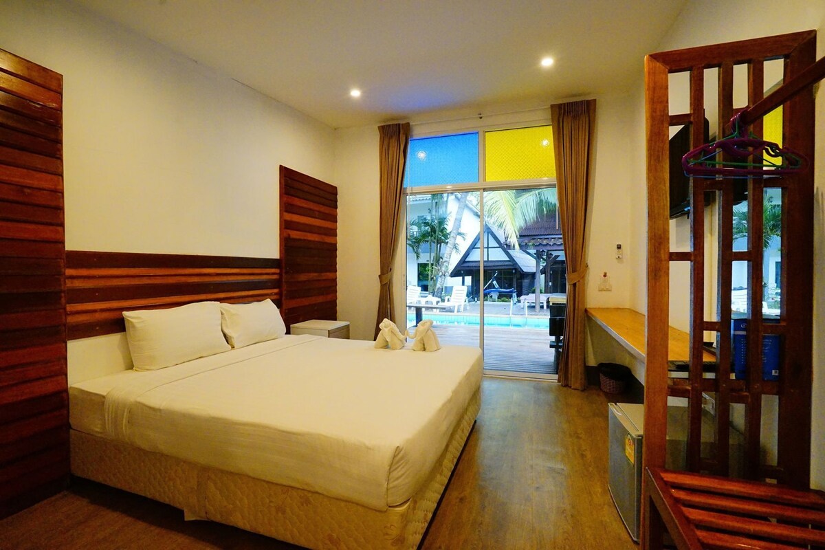 A hotel room with a large bed, wooden headboard, a small bedside table, and a view of a pool and building outside.