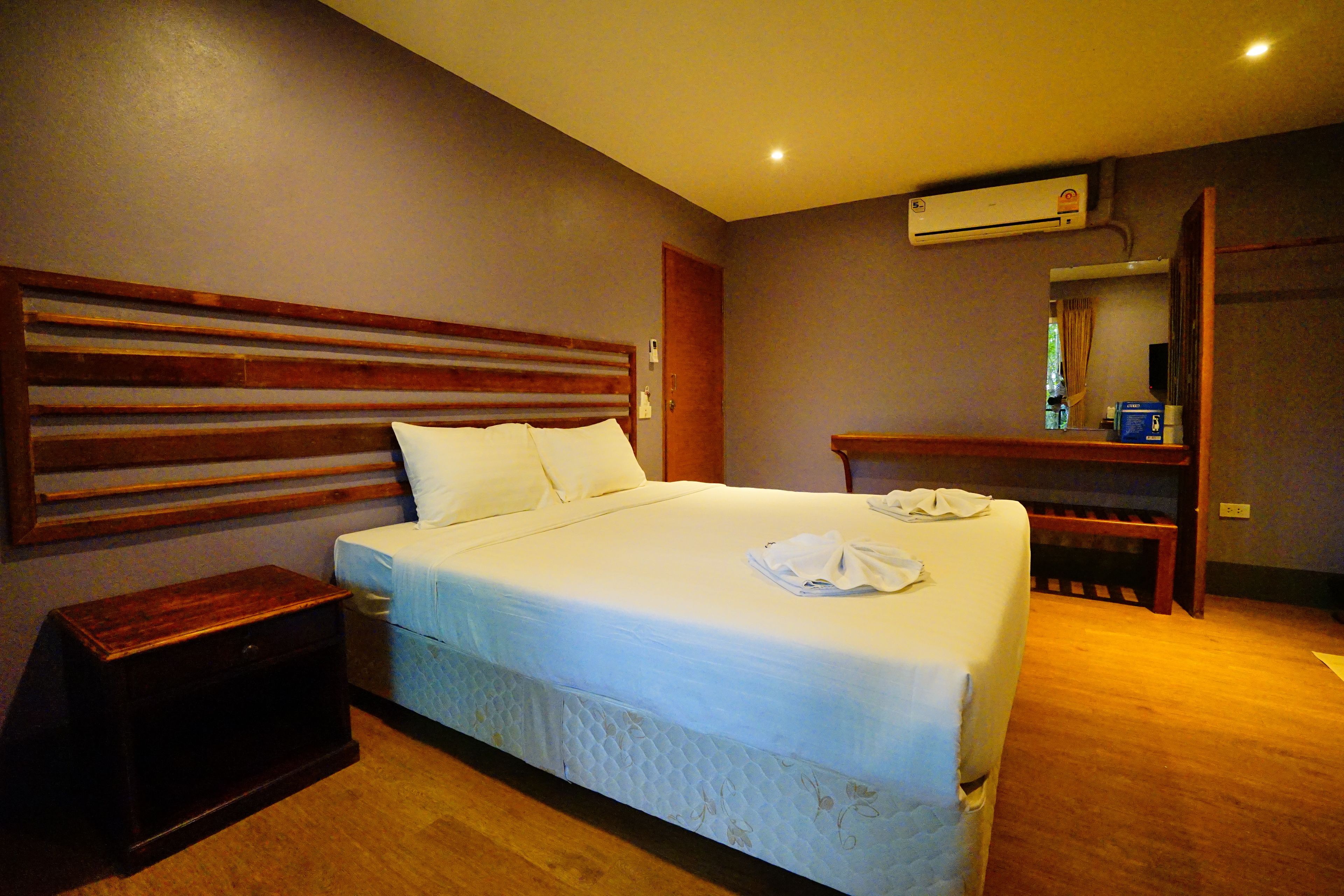 A hotel room with a large bed, a wooden headboard, a bedside table, an air conditioning unit, and a small bench.