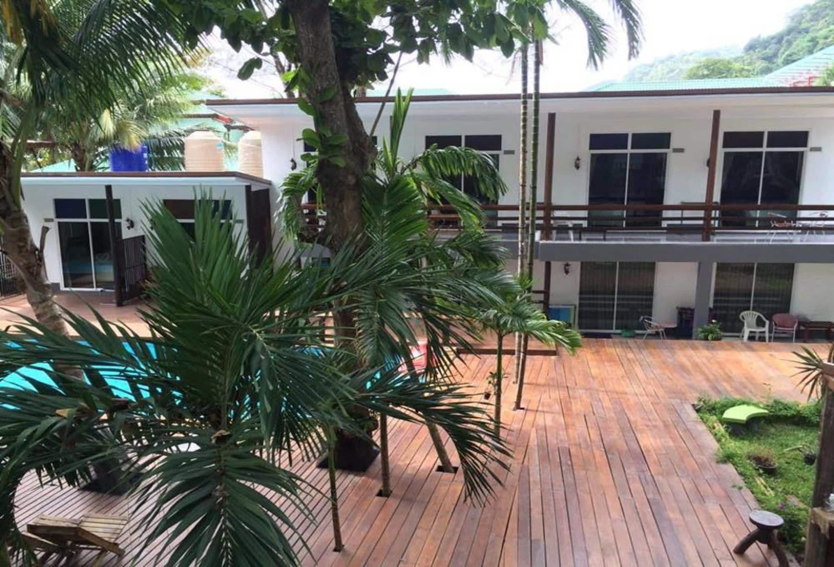 Una terraza de madera con palmeras, piscina y un edificio con balcón.