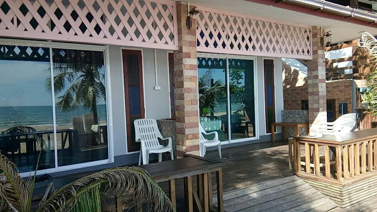 Uma área externa coberta com deck de madeira, cadeiras de plástico branco e uma mesa de madeira.