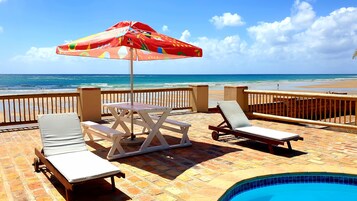 Un patio frente a la playa con piscina, sillas de descanso y una mesa con sombrilla.