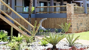A multi-story hotel with balconies, a palm tree, and a landscaped garden with various plants.