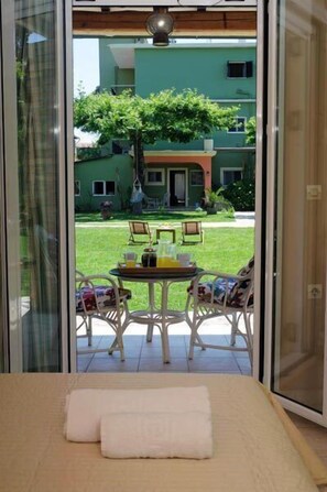 A room with a bed, a table with chairs, and a view of a green lawn and buildings outside. - Efi & Sofia Apartments (Corfu)