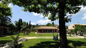 A courtyard with a building featuring a tiled roof, surrounded by lush greenery and trees. - Efi & Sofia Apartments (Corfu)