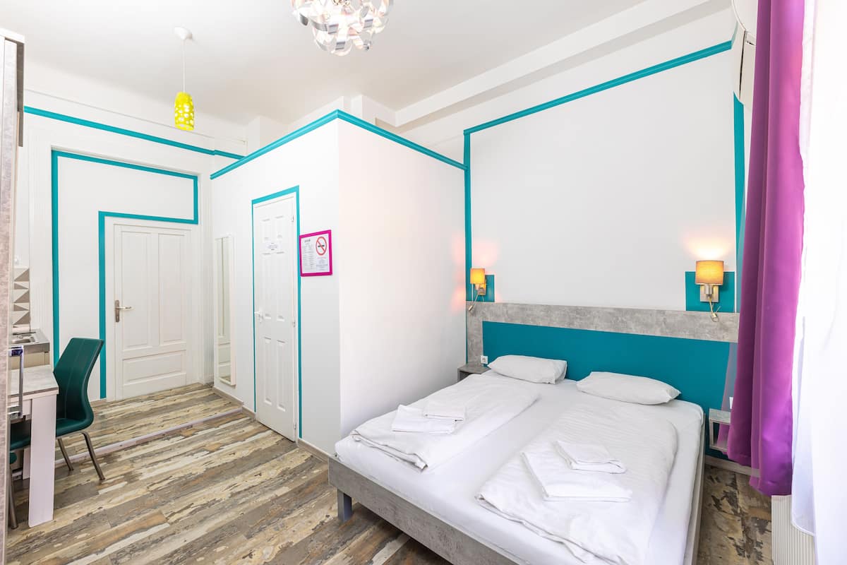 A hotel room with a bed, a desk, and a chair. There is a door and a wall with a sign.