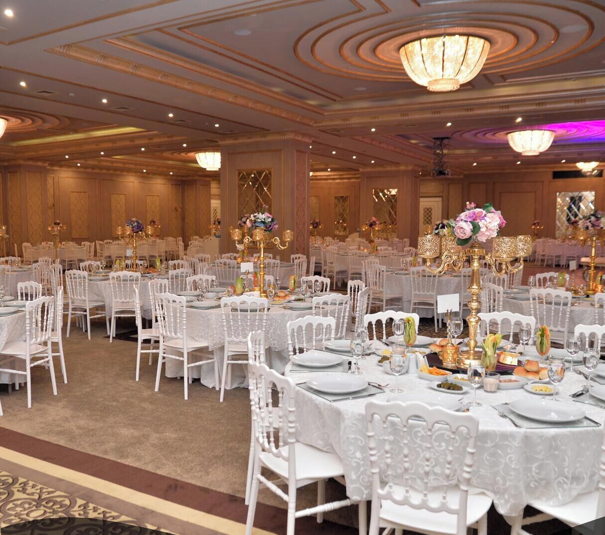 Un salón de banquetes con mesas redondas, sillas blancas y candelabros.