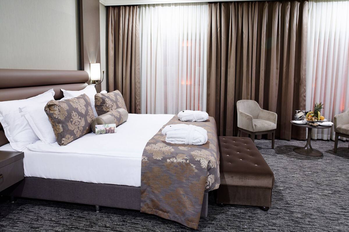 A hotel room with a large bed, two armchairs, a small table with fruit, and a view of the city through curtains.
