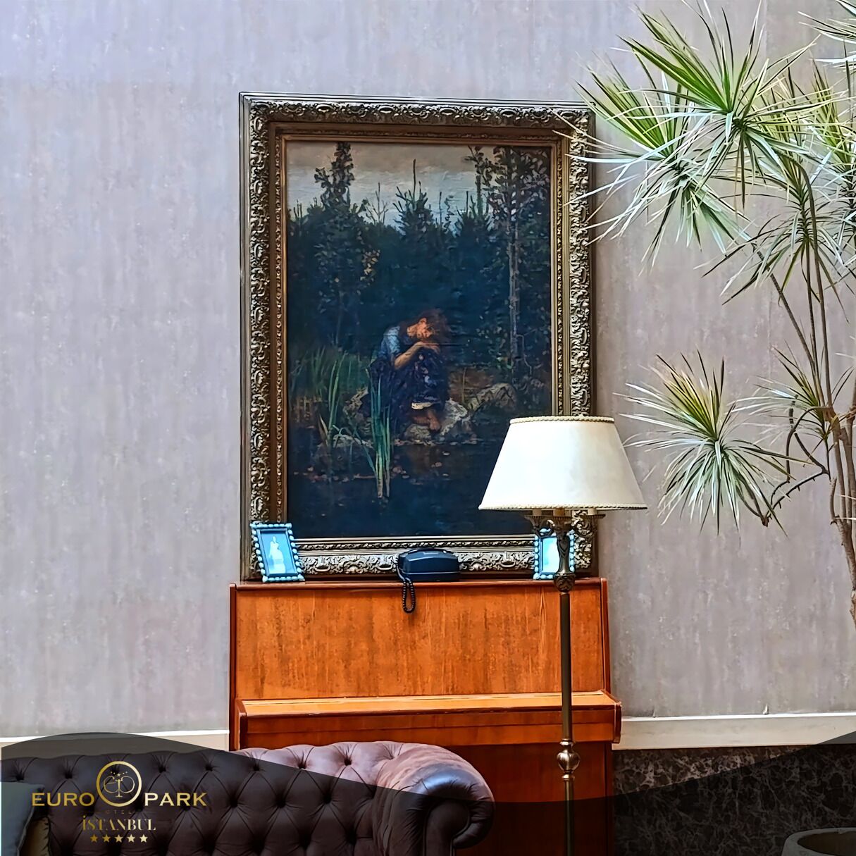 A framed painting of a person by a pond, a lamp, and a plant on a wooden table.