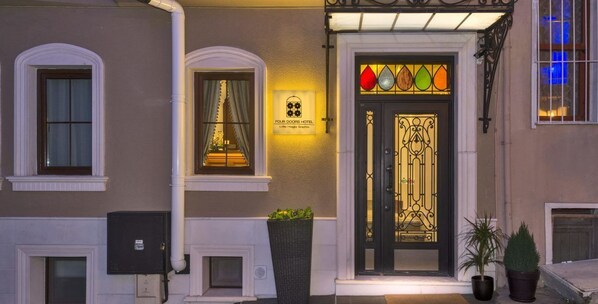 Entrada de hotel con una puerta negra decorativa, un letrero encima de la puerta y un pequeño ventanal con cortinas.
