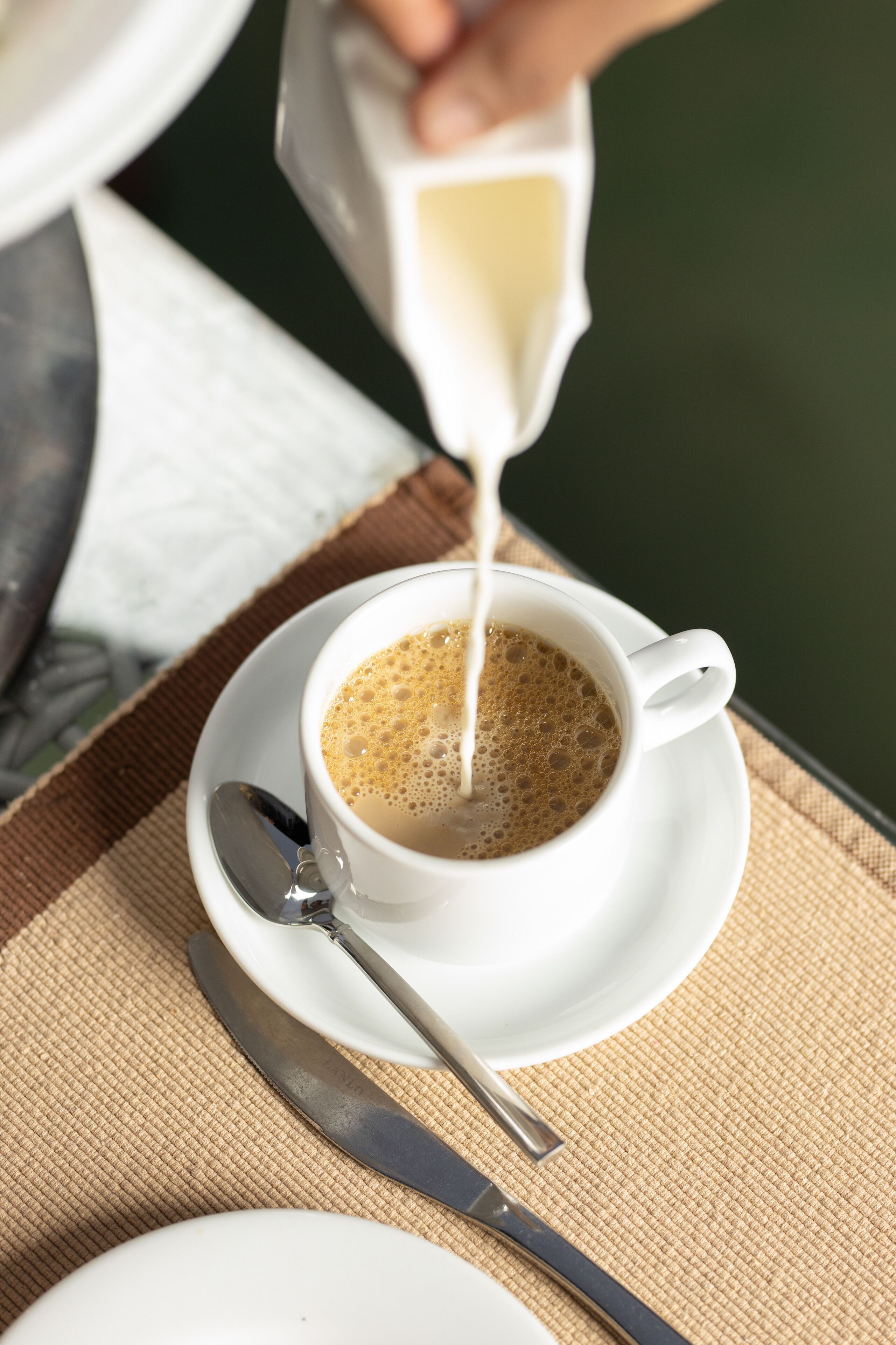 A cup of coffee with milk being poured into it, placed on a saucer with a spoon beside it.