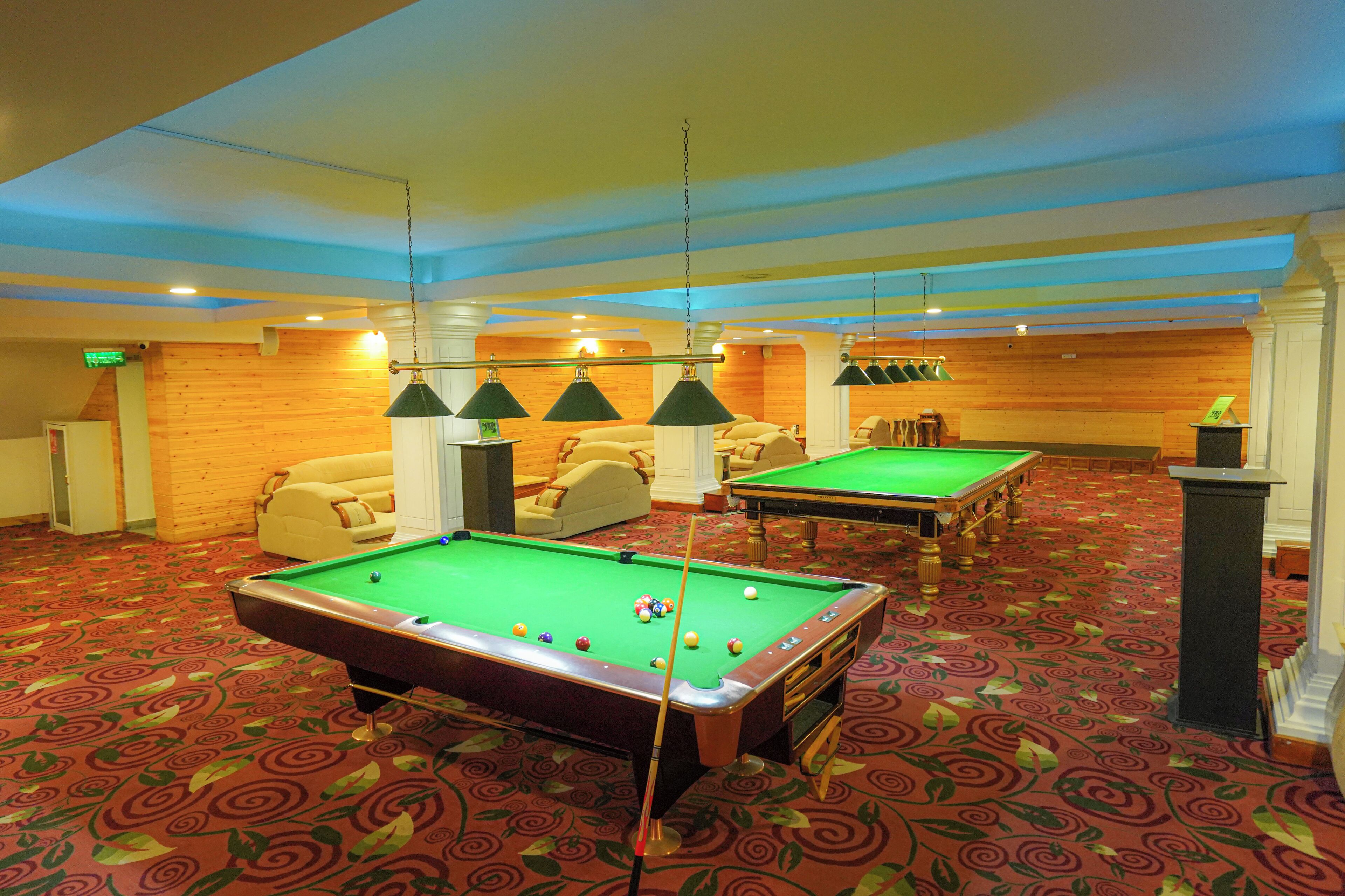 A pool table with balls, a sofa, and a billiard table in a room with patterned carpet and wooden walls.