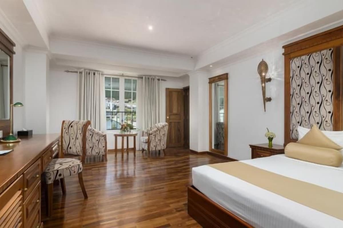 A hotel room with a wooden floor, a large bed with beige bedding, a desk with a lamp, two chairs, a small table with a vase, and a window with curtains.