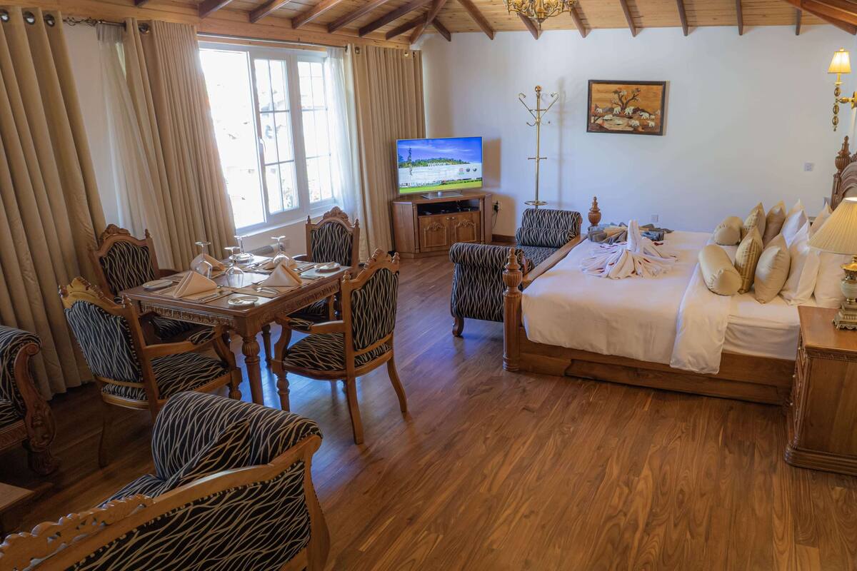 Ein Hotelzimmer mit Holzboden, einem Bett mit weißer Bettwäsche, einem Esstisch mit Stühlen, einem Fernseher und einem Gemälde an der Wand.
