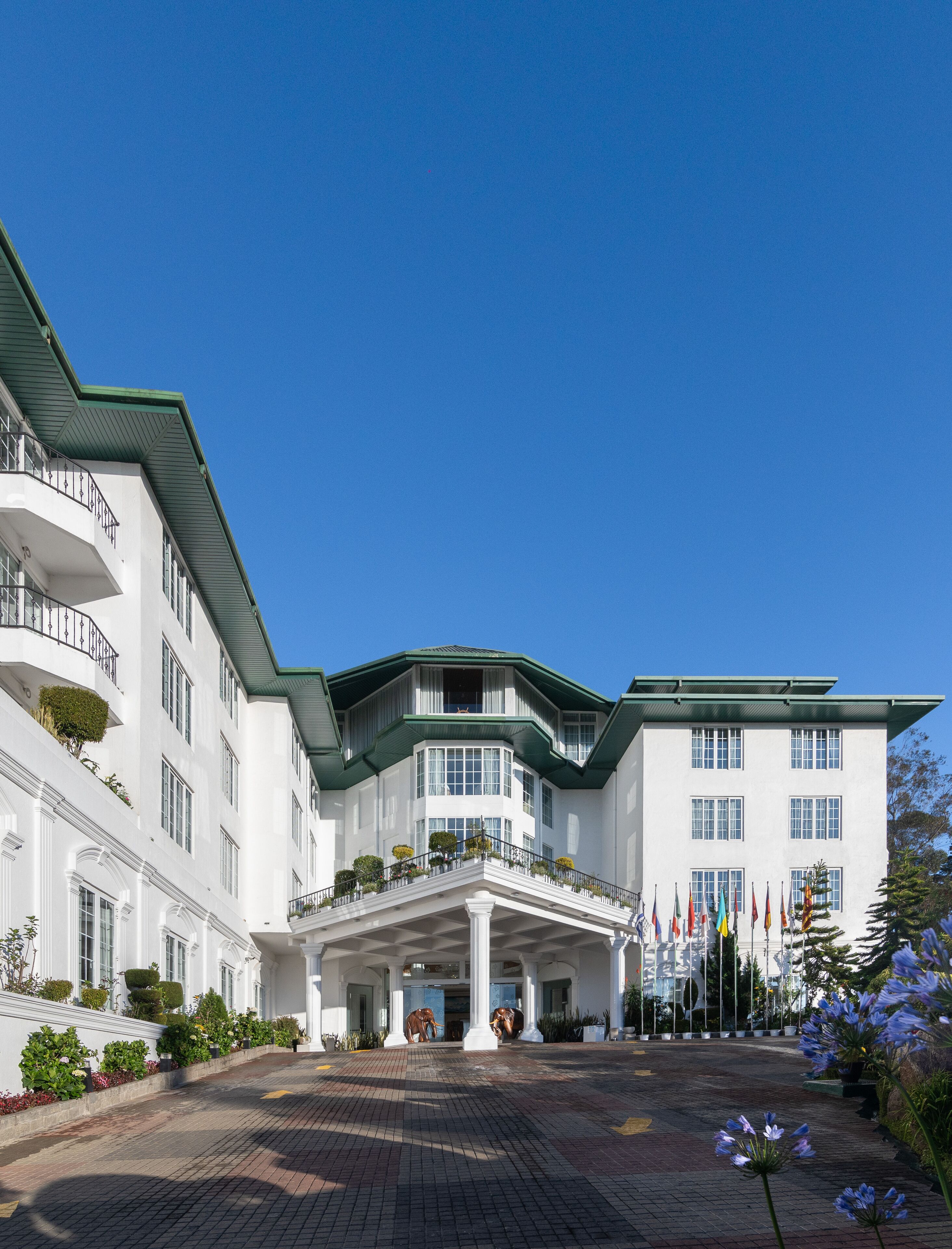 A large, white building with a green roof and multiple balconies. It features a central entrance with a covered walkway and several flags in front.