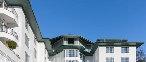 A large, white building with a green roof and multiple balconies. It features a central entrance with a covered walkway and several flags in front.