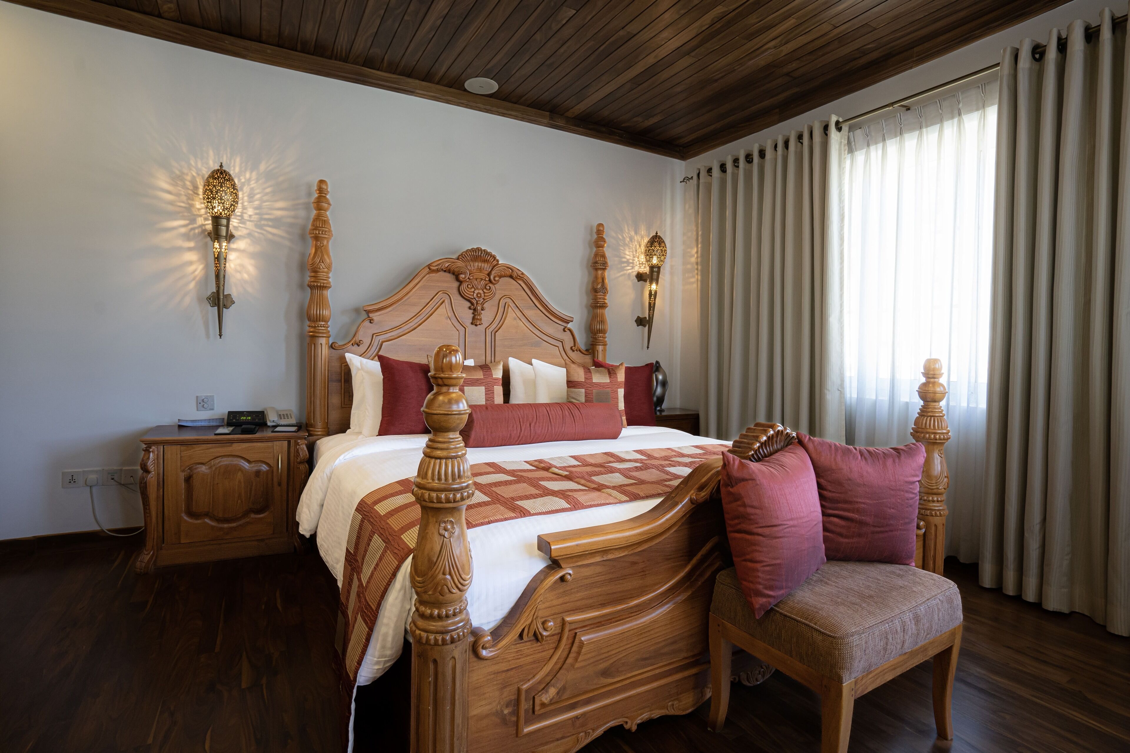 A wooden four-poster bed with a quilt and pillows, two bedside tables, a chair, and a window with curtains.