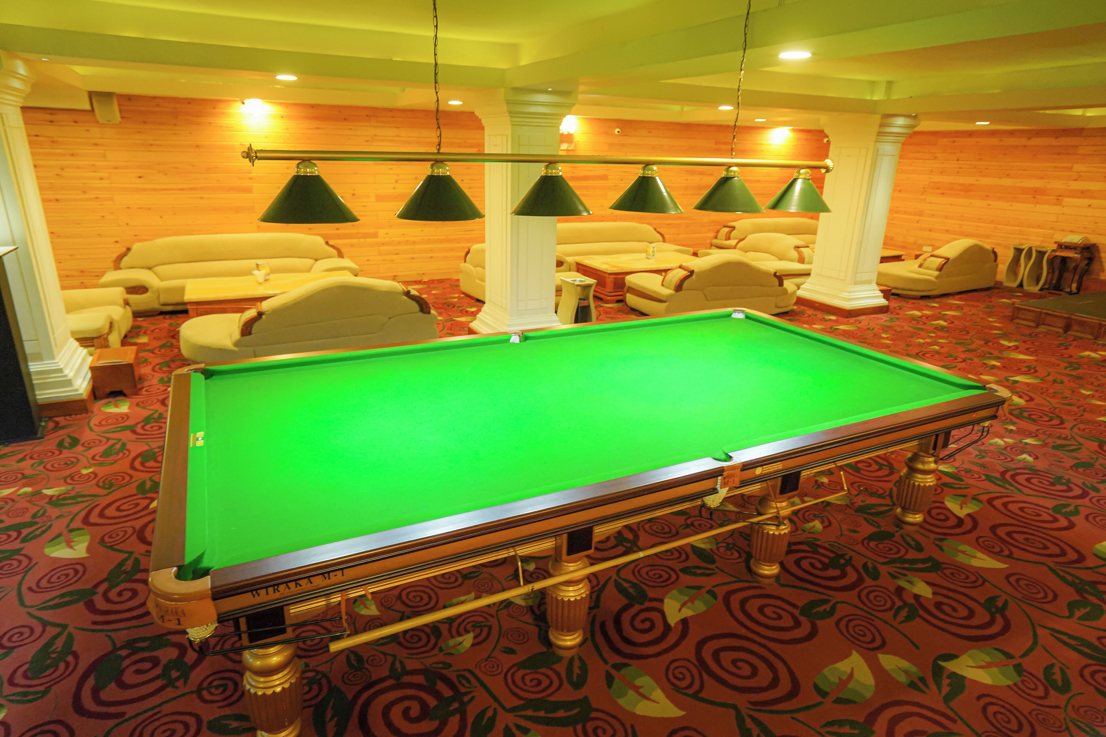 A pool table in a room with a red carpet, beige sofas, and green hanging lamps.